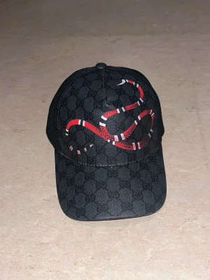 Gucci keps med orm och GG-mönster - Svart keps från Gucci med klassiskt GG-monogram över hela ytan och en röd-vit orm broderad framtill. Justerbar rem baktill med blå och röda ränder. Kepsen är tillverkad i canvas och mesh för en luftig känsla.