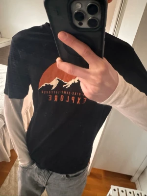 Svart t-shirt med tryck 'Explore' från Kappahl  - Säljer en svart t-shirt med orange och vitt tryck av berg och texten 'CLIMB HIKE CAMP DISCOVER EXPLORE'. T-shirten har en unik design med vita långa ärmar som ger en lager-på-lager-look. Perfekt för dig som gillar äventyrsstil. 