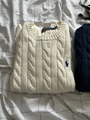 Vit kabelstickad tröja Polo Ralph Lauren - Vit kabelstickad tröja från Polo Ralph Lauren med rund halsringning och broderad marinblå logga på bröstet. Tröjan har långa ärmar och ribbstickade muddar samt nederkant. Perfekt för en klassisk och stilren look.