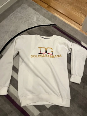 Vit sweatshirt från Dolce & Gabbana - Snygg vit sweatshirt från Dolce & Gabbana med broderad DG-logga och text i guld, svart och rött på bröstet. Tröjan har rund halsringning, långa ärmar och ribbade muddar vid ärmslut och nederkant. Perfekt för en clean och trendig look.