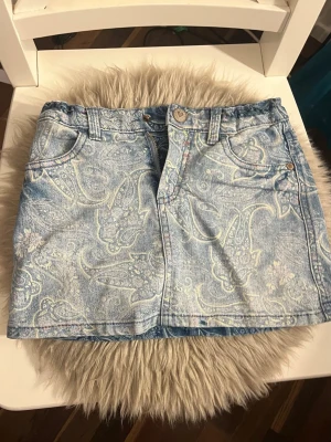 Ljusblå jeanskjol med paisleymönster - Säljer en ljusblå jeanskjol med snyggt paisleymönster. Kjolen har klassiska fem fickor, bälteshällor och knapp- samt dragkedjestängning framtill. Perfekt för dig som vill ha en unik twist på din denimstil.