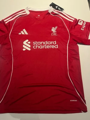 Liverpool FC matchtröja Adidas röd - Snygg röd Liverpool FC matchtröja från Adidas med vita detaljer och klubbmärke på bröstet. Tröjan har Standard Chartered som sponsortryck och är tillverkad i lätt och ventilerande material med AEROREADY-teknologi. Perfekt för fotboll eller träning.