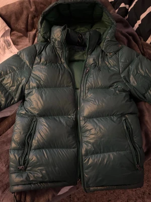 Ralph lauren i en snygg grön färg - Säljer en mörkgrön pufferjacka från Polo Ralph Lauren. Jackan har huva, två stora fickor med dragkedja och en hög krage. Materialet är glansigt och känns riktigt lyxigt. Perfekt för kalla dagar när du vill hålla stilen. Säljs pga att den är för liten. Köpt 2024 tror jag och är i fint skick! Minsta funderingar så hör av er!