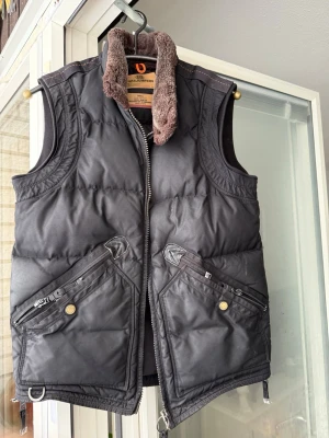 Svart dunväst från Parajumpers - Säljer en svart dunväst från Parajumpers med brunt fuskpälsfoder i kragen, stora snedställda fickor med dragkedja och detaljer i metall. Västen har quiltad design och robust dragkedja framtill. Perfekt för kalla dagar och riktigt snygg till lager-på-lager.