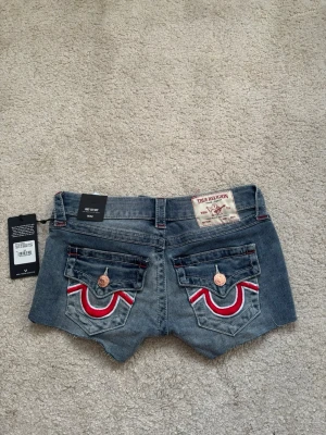 True Religion denimshorts blå W24 - Säljer ett par  blå denimshorts från True Religion med snygga kontrastsömmar och broderade fickor bak i rött och vitt. Låg midja och är stretchiga med prislapp kvar.