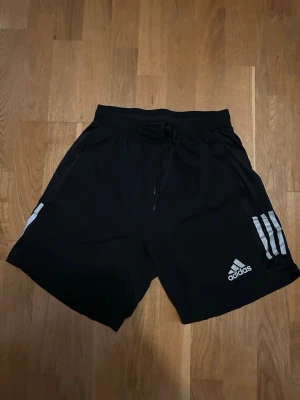 Svarta Adidas träningsshorts - Säljer ett par svarta träningsshorts från Adidas med klassiska vita ränder på sidorna och logga framtill. Shortsen har snörning i midjan och är tillverkade i ett lätt och bekvämt syntetmaterial, perfekta för sport och aktivitet.