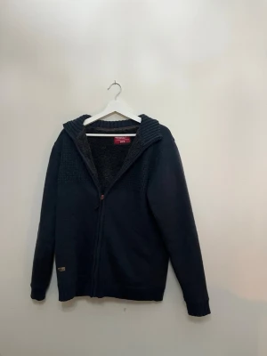 Mörkgrå stickad kofta med fleecefoder - Mysig mörkgrå stickad kofta från Hero med dragkedja och fluffigt fleecefoder på insidan. Koftan har ribbad krage och muddar samt diskret märke nertill. Perfekt för kalla dagar när du vill vara både varm och stilren.