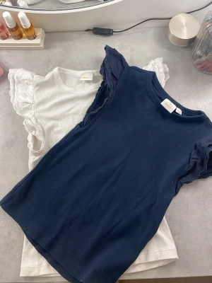 Volangärmad topp Gina Tricot 158/164 - Två ribbade toppar från Gina Tricot i storlek 158/164. En marinblå och en vit, båda med volangprydda korta ärmar i broderad bomull. Perfekta för dig som vill ha en söt och enkel look med extra detalj på axlarna.💙🤍 Man kan såklart köpa det separat också. Det kostar 70kr styck. 