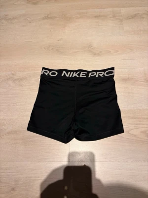 Nike Pro svarta träningstights shorts - Säljer två par svarta Nike Pro shorts i tight modell, perfekta för träning. Ena paret har bred resår med vit Nike Pro-logga, det andra har både svart och röd resår samt Nike-märke och swoosh i rött och vitt på benet. Stretchigt material som sitter skönt och följsamt.