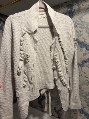 Vit stickad kofta med brodyr - Säljer en chunky vit stickad kofta från Odd Molly med broderad patch på ärmen, volangdetaljer framtill och knytband i midjan. Koftan har lång ärm och en cool struktur som gör den extra mysig och unik.