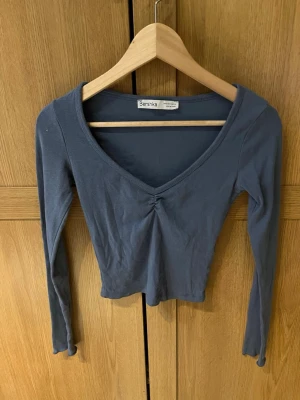 Mörkblå croppad topp från Bershka - Snygg mörkblå croppad topp från Bershka med lång ärm och v-ringning. Toppen har en liten rynkning framtill vid bysten och är tillverkad i mjuk bomullsmix. Perfekt till jeans eller kjol för en enkel men trendig look.