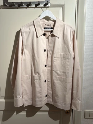 Beige overshirt från J.Lindeberg - Stilren beige overshirt från J.Lindeberg med stora framfickor och svarta knappar. Klassisk krage och rak passform, perfekt att bära över en t-shirt. Tillverkad i mjuk bomull för en clean och modern look. Helt oanvänd med prislappen kvar.