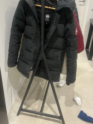 Svart dunjacka från Canada Goose XS - Säljer en svart, lång dunjacka från Canada Goose i storlek XS. Jackan har klassisk patch på ärmen, dragkedja och tryckknappar framtill. Fylld med dun för extra värme och har en stilren, quiltad design. Perfekt för kalla vinterdagar.