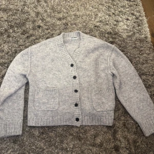 Ljusgrå kofta från Weekday - Mysig ljusgrå kofta från Weekday med v-ringad hals, stora framfickor och svarta knappar. Stickad i mjukt material och har ribbade muddar vid ärmslut och nederkant. Perfekt för lager på lager under kyliga dagar.