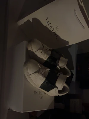 Valentino Open sneakers vit/svart - Säljer ett par Valentino Open sneakers i vitt skinn med bred svart rem över mitten. Klassisk chunky sula och rund tå. Skorna har snörning och subtila nitar bak på hälen för extra edge. Perfekta för dig som vill ha en clean men ändå statement look.