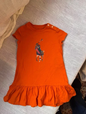 Orange barn klänning från Ralph Lauren - Superfin orange barn klänning från Ralph Lauren i mjuk bomull. Klänningen har korta ärmar, volang nertill och en stor färgglad Polo-logga framtill. 