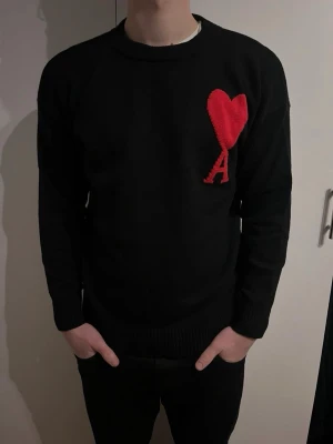 Svart stickad tröja Ami Paris hjärta - Svart stickad tröja från Ami Paris med ikoniskt rött hjärta. Stilren tröja med en väldigt snygg look.