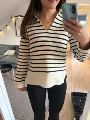 Randig vit tröja med krage - Säljer en vit långärmad tröja med svarta horisontella ränder och klassisk krage. Tröjan har v-ringad öppning framtill och är tillverkad i ett mjukt stickat material. Passformen är normal och den har små slitsar i sidorna för en avslappnad look.