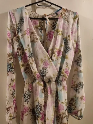 Blommig omlotttopp från Blood+Bone - Supersöt omlottopp från Blood+Bone med blommigt mönster i pastelliga färger. Toppen har lång ärm, v-ringning och knyt i ryggen. Tunt, luftigt tyg med innerfoder och resår i midjan för en snygg passform.