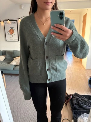 Grön stickad kofta med knappar - Mysig grön stickad kofta med v-ringning och stora framfickor. Koftan har ribbade muddar, knäppning med fyra knappar och en lite loose passform. Perfekt för lager på lager under höst och vinter.