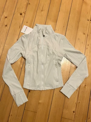 Lululemon Define Cropped Jacket  - Säljer en helt ny offwhite Define Cropped Jacket från Lululemon med figurnära passform och hel dragkedja framtill. Fick den i julklapp men var för liten och kunde inte skickas tillbaka då det gått för lång tid. Jackan har tumhål vid ärmsluten och diskret logga på ryggen. Perfekt för träning eller som ett snyggt lager till vardags. Tillverkad i stretchigt syntetmaterial. Nypris: 1380 kr