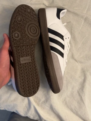 Adidas Samba sneakers vit/svart - Klassiska Adidas Samba sneakers i vitt skinn med svarta ränder och brun sula. Tåpartiet är i grå mocka och skorna har snörning. Guldtexten 'SAMBA' på sidan ger en retrovibe. Perfekta för dig som gillar tidlös streetstyle.