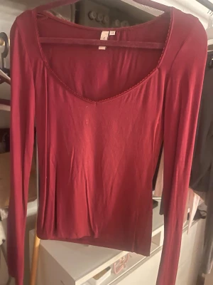 Mörkröd långärmad topp nelly - En enkel och snygg mörkröd långärmad topp från nelly. Toppen har en djup rundringning med dekorativ kant och är gjord i ett mjukt, stretchigt material. Perfekt att styla med jeans eller kjol för en clean look.