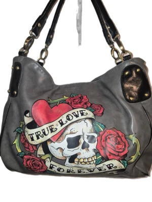Superfin stor axelväska med döskalle och rosor Ed Hardy - Cool grå axelväska från Ed Hardy med stort tryck av döskalle, hjärta och röda rosor samt texten 'True Love Forever'. Väskan har svarta handtag, guldfärgade detaljer och är gjord i skinnimitation. Insidan har flera fack och dragkedja. Perfekt för dig som gillar edgy stil!                                                                                    Som ny!                                                                            Djur och rökfritt hem.                                 