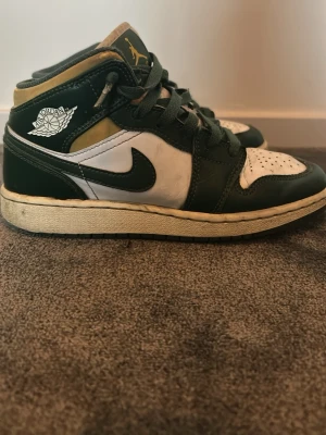 Nike Air Jordan 1 svart/vit/guld - Säljer ett par Nike Air Jordan 1 med svart, vit och gulddetaljer. Klassisk high-top modell med snörning och ikoniska Air Jordan-loggan på sidan. Skorna har en rund tå och är tillverkade i skinn med kontrasterande färgblock.