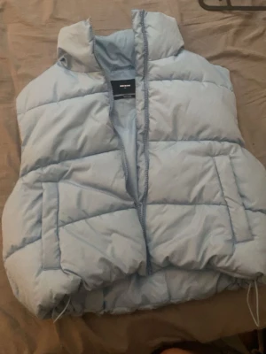 Ljusblå dunväst från Bik Bok - Säljer en ljusblå dunväst från Bik Bok i storlek M. Västen har hög krage, dragkedja framtill och två fickor på sidorna. Perfekt att slänga över en hoodie för en chill look. Tillverkad i återvunnet material.