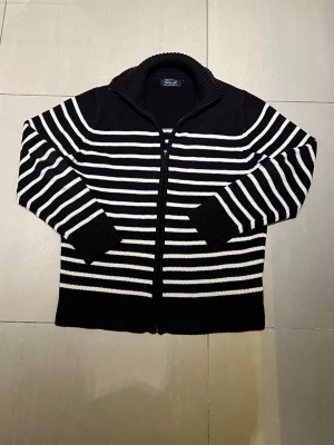 Svartvit randig kofta från Skill - Säljer en svart och vit randig kofta från Skill med dragkedja framtill och hög krage. Tröjan är stickad i tjockt material och har långa ärmar med breda muddar. Perfekt för kyliga dagar när du vill vara både bekväm och stilren.