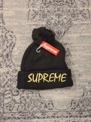 Supreme-New Era mössa - Supreme mössa i svart ribbstickat material med stor tofs på toppen. Broderad Supreme-logga i gult framtill och liten svartvit Supreme-tag baktill. Klassisk uppvikt kant och riktigt streetwear-vibe. Skriv vid frågor, priset är förhandlingsbart vid snabb affär och kan skicka direkt efter köpet 😀