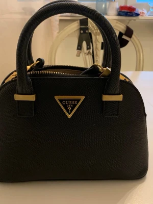 Lossie Dome Guess handväska - Snygg svart handväska från Guess med major gold detaljer och klassisk trekantslogga framtill. Väskan har två rejäla handtag, justerbar axelrem och dragkedja upptill. Materialet är strukturerat syntet som ger en exklusiv känsla. Helt ny Guess handväska. Tillkommer dustbag   Passform:  Höjd: 18,5 cm i storlek one size  Längd: 27 cm i storlek one size  Vidd: 29,5 cm i storlek one size.
