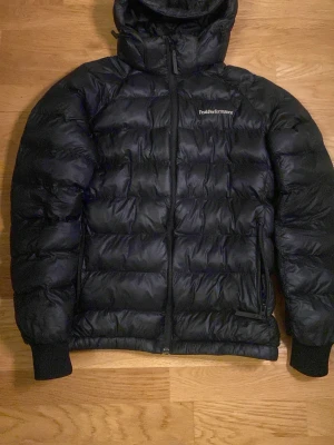 Svart dunjacka Peak Performance 160 - Säljer en svart dunjacka från Peak Performance med huva och dragkedja. Jackan är quiltad och har ribbade muddar vid ärmsluten. Klassisk logga på bröstet och en diskret detalj på ärmen. Perfekt för kalla dagar och riktigt snygg passform.