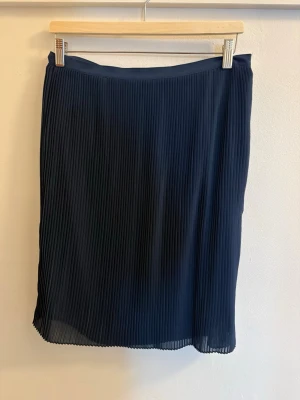 Mörkblå plisserad kjol - Säljer en mörkblå plisserad kjol med snygga smala veck. Kjolen har en rak passform och slutar strax ovanför knät. Tillverkad i ett lätt och luftigt syntetmaterial som ger ett fint fall. Perfekt att matcha med en enkel topp eller stickad tröja.