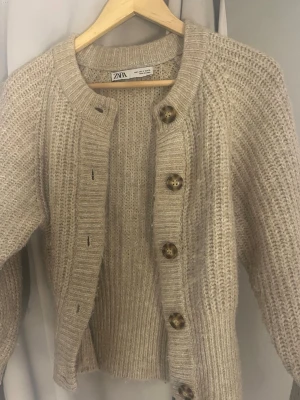 Beige stickad kofta från Zara - Mysig beige stickad kofta från Zara med stora bruna knappar framtill. Koftan har en  ribbad struktur, rund halsringning och långa ärma.
