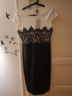 Svartvit spetsdetaljerad klänning - Snygg svartvit klänning med korta ärmar och spetsbroderier i svart över bysten. Klänningen har en figurnära passform och är knälång. Överdelen är vit och nederdelen svart, vilket ger en klassisk och elegant look.