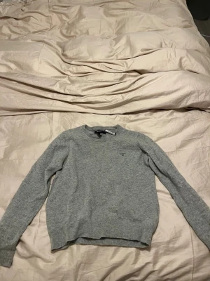 Grå lammullströja från GANT - Säljer en stilren grå tröja från GANT i mjuk lammull. Tröjan har rund halsringning, ribbade muddar och diskret GANT-brodyr på bröstet. Perfekt för lager-på-lager under kyliga dagar.