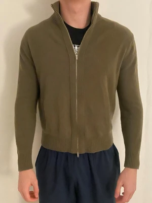 Olivgrön kofta från Massimo Dutti - Olivgrön kofta från Massimo Dutti i storlek L men passar även M. Tröjan har hel dragkedja framtill, ribbade muddar och hög krage. Tillverkad i mjukt stickat material, perfekt för lager-på-lager under kyliga dagar.