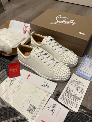 Vita sneakers från Christian Louboutin - Säljer ett par vita sneakers från Christian Louboutin i skinn med ikoniska nitar på tån. Skorna har rund tå, snörning och den klassiska röda sulan. Perfekt för dig som vill sticka ut med en lyxig och edgy stil. Kommer med originalkartong och certifikat.