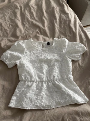Vit spetsblus från SHEIN - Supersöt vit blus från SHEIN med puffärmar och peplumdetalj i midjan. Blusen har ett vackert spetsmönster och rund halsringning. Perfekt att styla med jeans eller kjol för en fräsch look. Stängs med knapp i nacken.