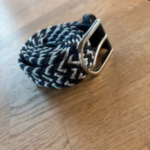 Randigt flätat bälte med silverfärgat spänne - Snyggt flätat bälte med randigt mönster i marinblått, vitt och brunt. Bältet har ett stilrent silverfärgat metallspänne och detaljer i mörkblått skinn vid ändarna. Perfekt för att ge outfiten lite extra edge.