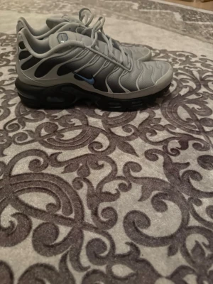 Nike Air Max Plus Tn grå och svarta - Nike Air Max Plus Tn sneakers i grått och svart med blå detaljer och Tn-logga bak. Skorna har vågiga linjer på ovandelen, svart sula med synliga luftbubblor och snörning. Snygg och sportig modell som sticker ut med sin unika design och coola färgkombination. ÄKTA