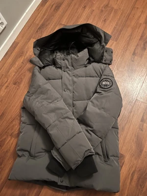 Grå dunjacka från Canada Goose - Säljer en grå dunjacka från Canada Goose med avtagbar huva och snygg pälskant. Jackan har patch på ärmen, dragkedja och knappar framtill samt två fickor. Perfekt för kalla dagar och riktigt varm. Passar dig som vill ha en stilren och funktionell vinterjacka.