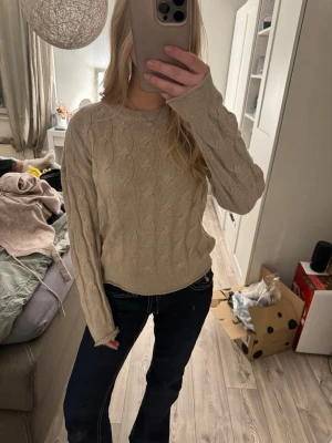 Beige kabelstickad tröja - Säljer en beige kabelstickad tröja med rund hals och långa ärmar. Tröjan har ett klassiskt flätmönster och är i mjukt material som passar perfekt till jeans eller kjol. Enkel och snygg att styla till olika outfits.