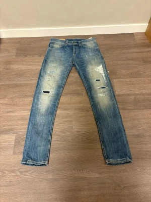 Blå slitna jeans från Dondup - Säljer ett par blå jeans från Dondup med coola slitningar och hål på benen. Jeansen har klassisk femficksdesign, normal passform och är tillverkade i bomull. Perfekta för dig som gillar en avslappnad och trendig look.