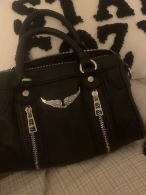 Zadig & Voltaire väska - Säljer en svart handväska i skinn med coola silverfärgade dragkedjor och ett snyggt emblem med glittrande vingar framtill. Väskan har dubbla handtag och axelrem, perfekt storlek för det viktigaste. Stilren och edgy design som lyfter vilken outfit som helst. Säljer då jag ej använder längre och har flera liknande, vet ej om den är äkta eller inte men ganska säker på att den är de men kan inte lova något då jag inte vet till 100%💕