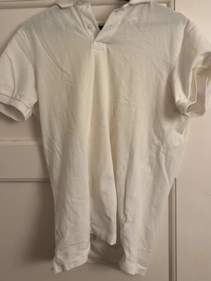 Vit pikétröja från Dressmann M - Klassisk vit pikétröja från Dressmann i storlek M. Tröjan har korta ärmar, krage och två knappar vid halsen. Tillverkad i bomull för en skön och luftig känsla. Perfekt till en clean och enkel stil.