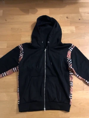 Burberry hoodie med dragkedja – svart, nytt skick. - Säljer en Burberry zip hoodie/kofta i svart med klassiska Burberry-rutiga sidopaneler. Storlek: S Skick: nytt skick, inga hål eller fläckar. Använd sparsamt. Säljs pga passform. Skickas spårbart eller möts upp.