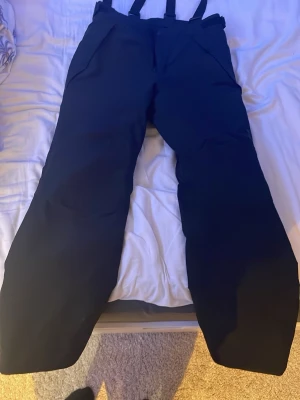 Svarta täckbyxor från Everest - Svarta täckbyxor från Everest med justerbara hängslen och dragkedja fram. Byxorna är tillverkade i slitstarkt syntetmaterial och har en loose passform som ger bra rörelsefrihet. Perfekta för kalla dagar i backen eller snölek.